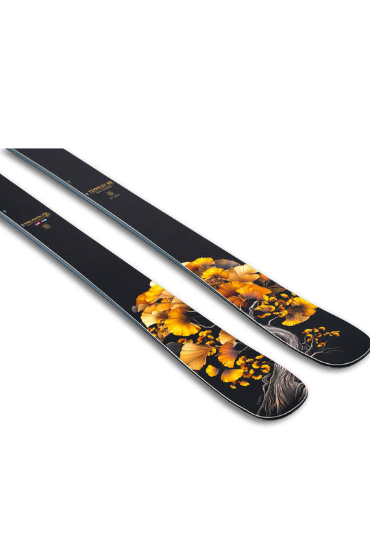 A pair of black skis with a detailed graphic print of golden autumn leaves and tree branches, displayed diagonally on a clean white background.