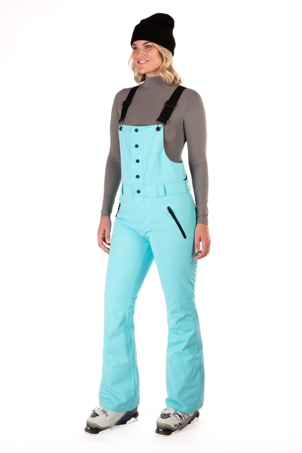 A woman wearing a blue snow bib over a gray shirt with black straps, paired with light blue snow pants and gray boots, accessorized with a black beanie.