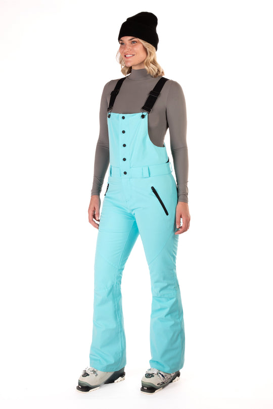 A woman wearing a blue snow bib over a gray shirt with black straps, paired with light blue snow pants and gray boots, accessorized with a black beanie.