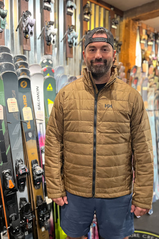 Man wearing a brown jacket in a ski shop with skis and equipment in the background