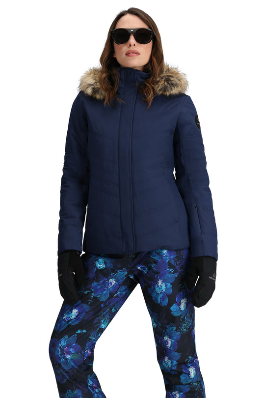 Woman wearing a blue ski jacket with fur hood, sunglasses, and floral pants on a white background
