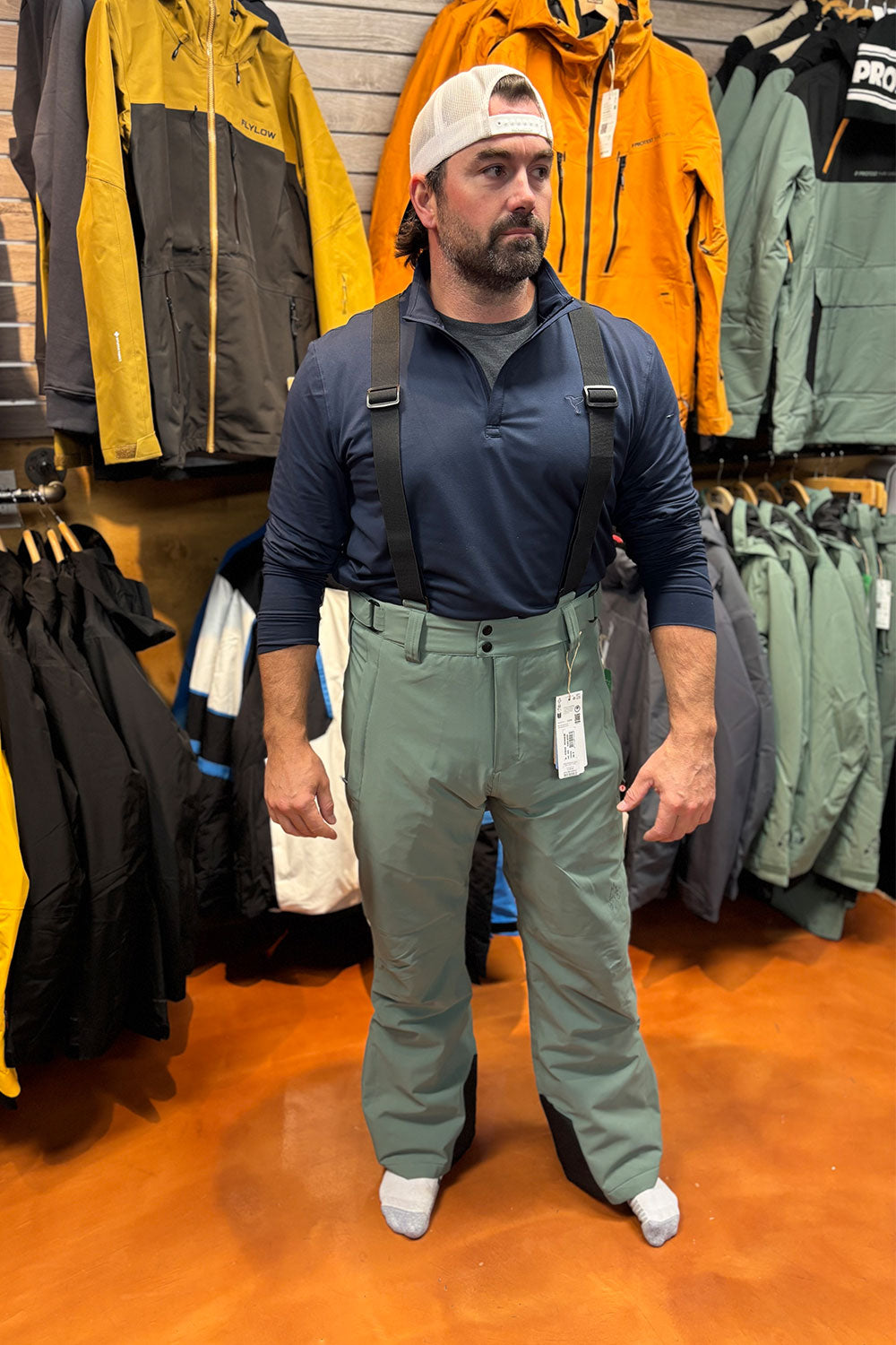 Man wearing a blue shirt, green pants, and a white cap standing in a clothing store with jackets on display.