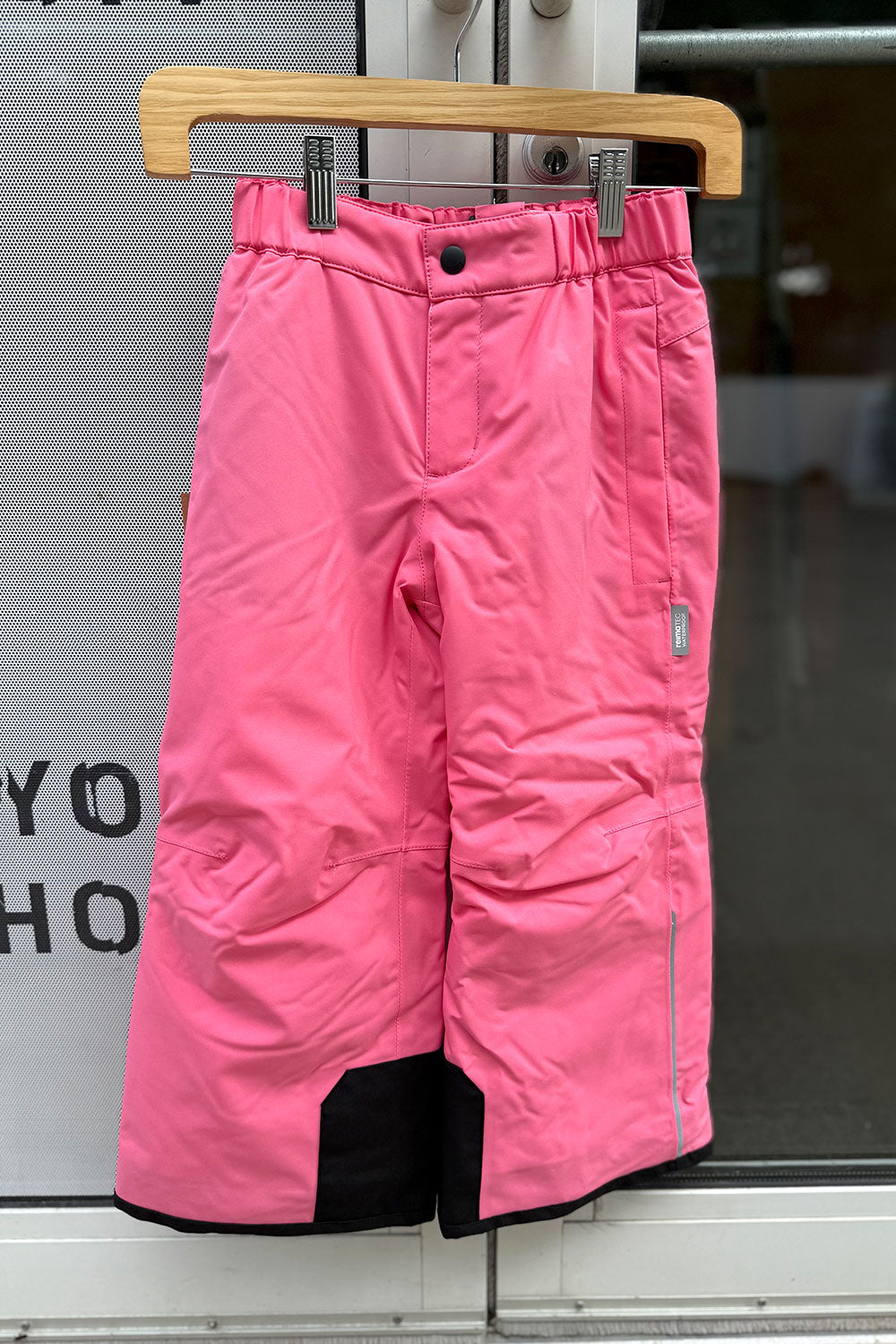 Pink snow pants hanging on a wooden hanger against a glass door background.