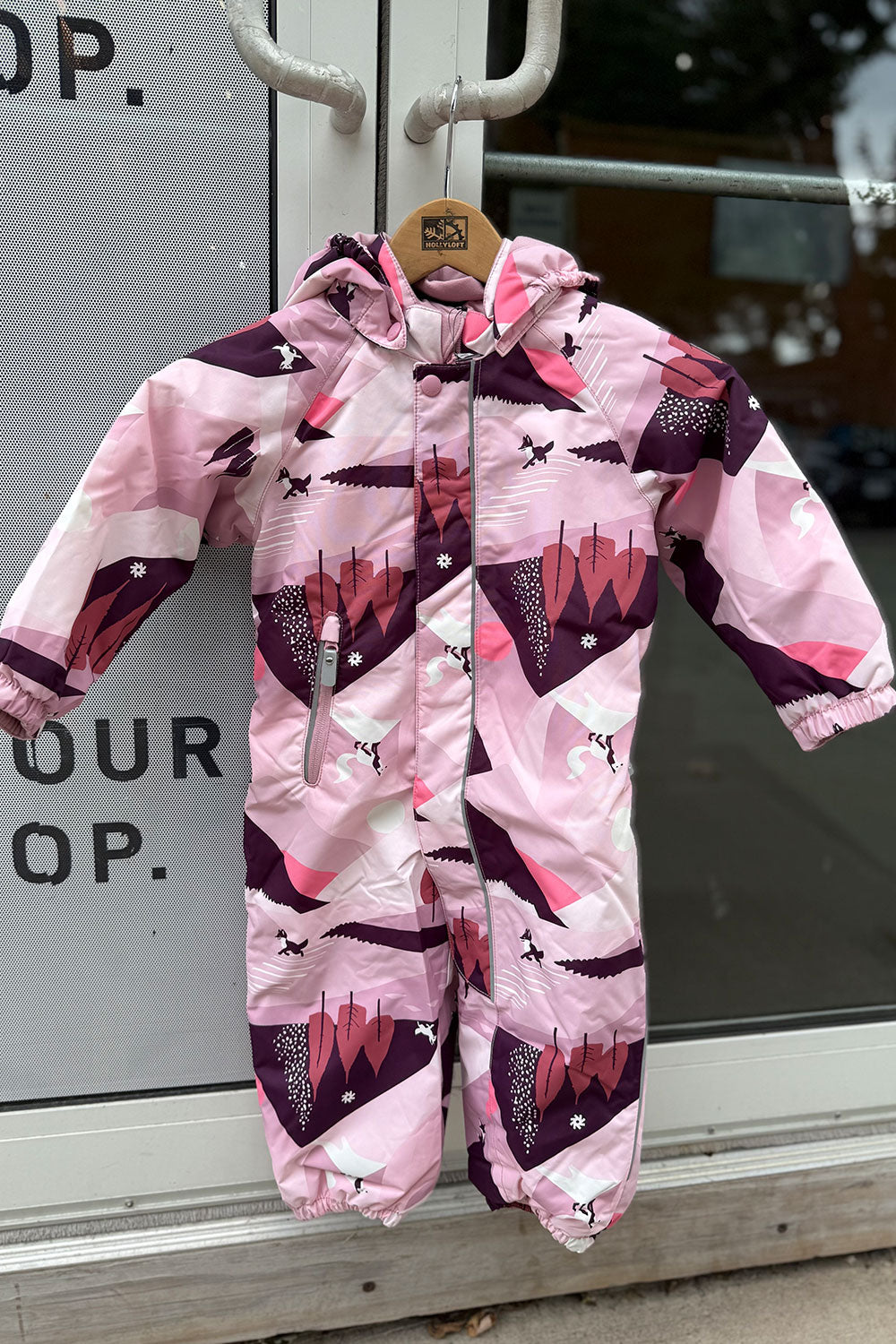 Pink and purple children's snowsuit with geometric pattern on a hanger against a glass door background.