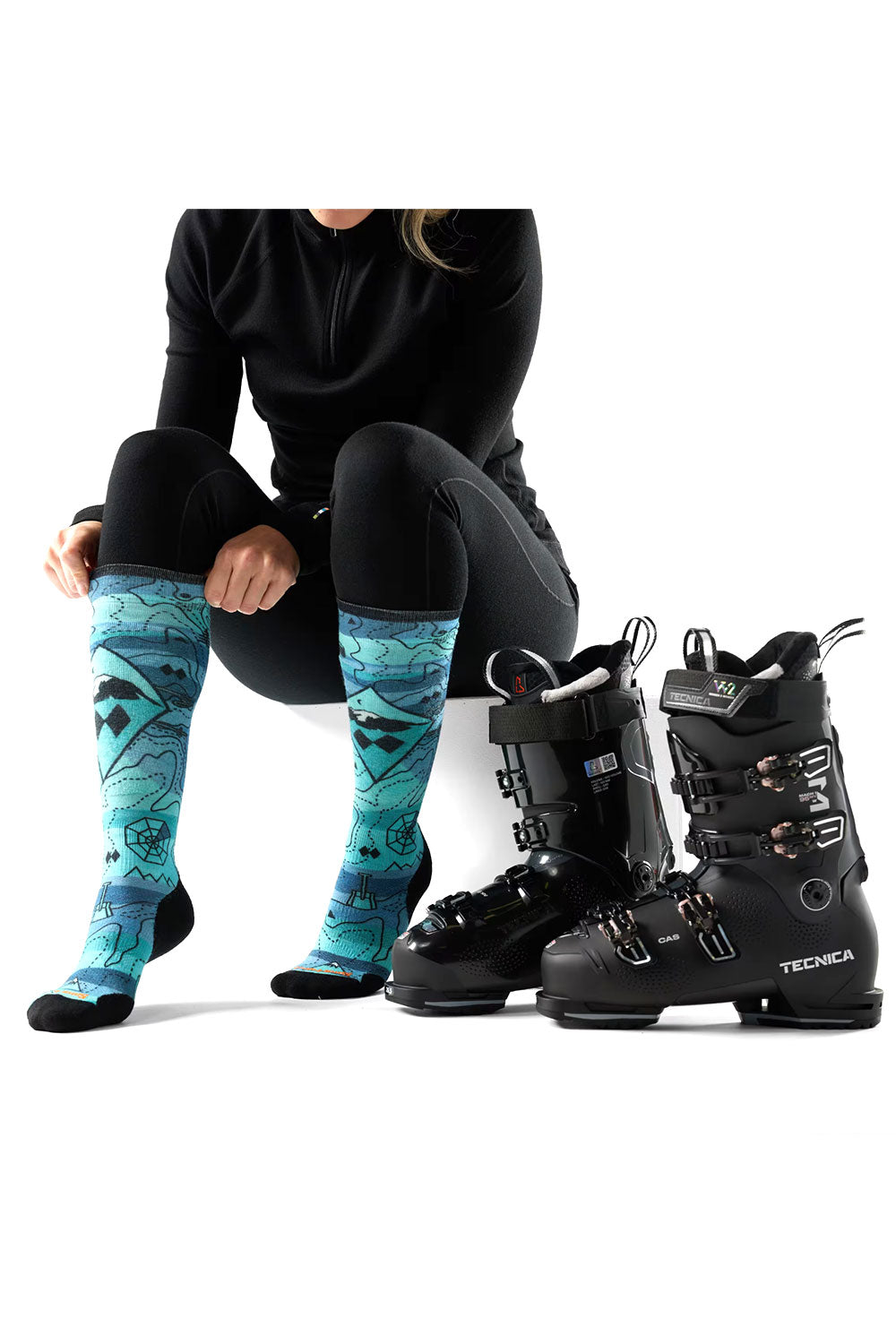 Person wearing black ski boots and blue patterned socks sitting next to a pair of black ski boots on a white background.
