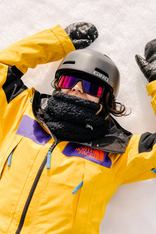 Person wearing a yellow North Face jacket, gray helmet, black neck gaiter and black gloves in the snow.