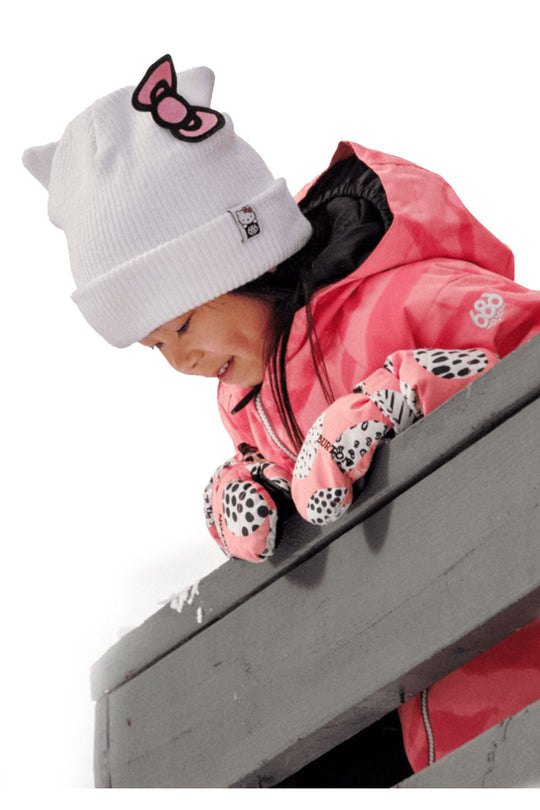 Child wearing a pink snowsuit with a white beanie and gloves, standing on a concrete surface.