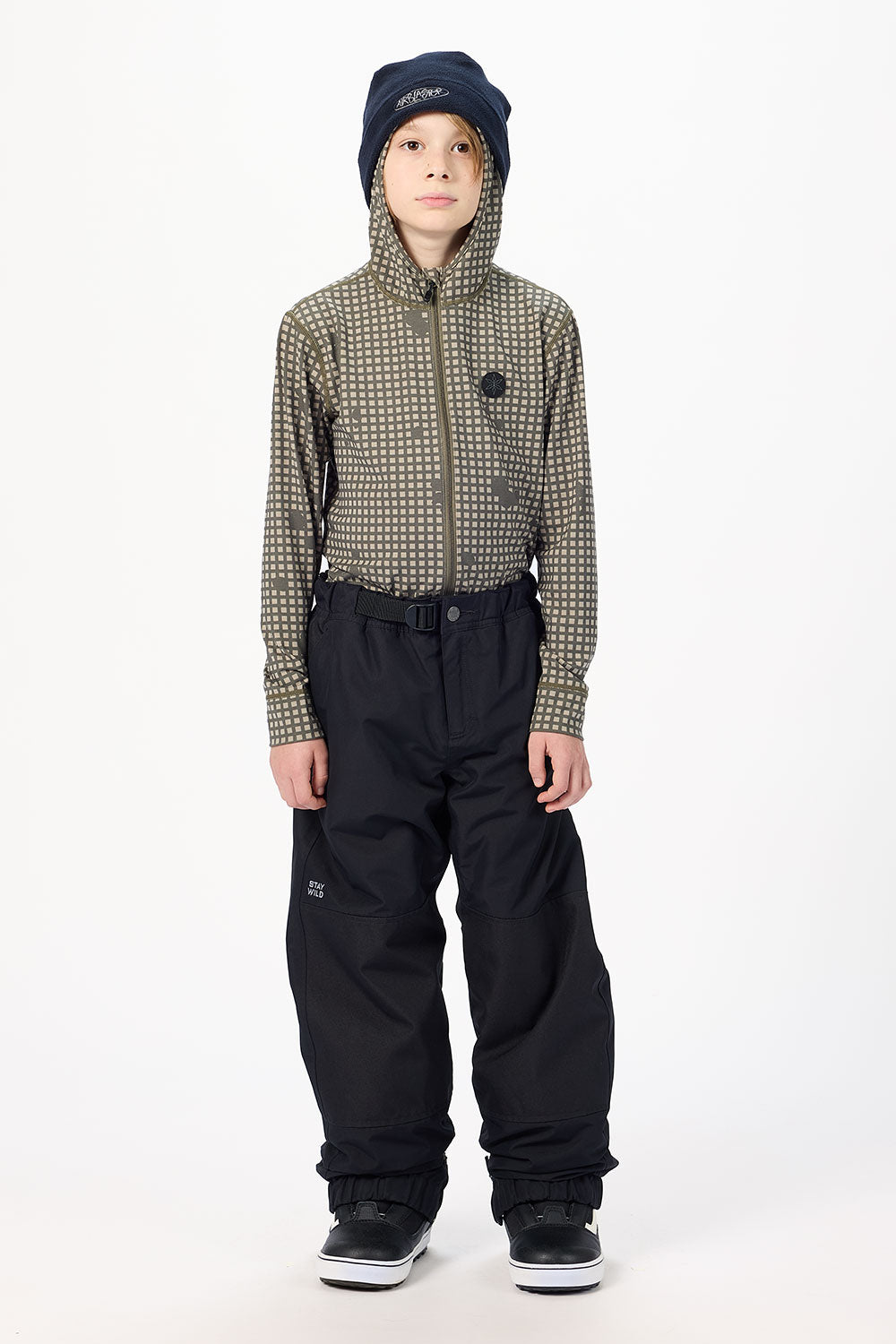A child model wearing a patterned zip-up jacket, black snow pants, a navy beanie, and black snow shoes, standing against a plain white studio background.