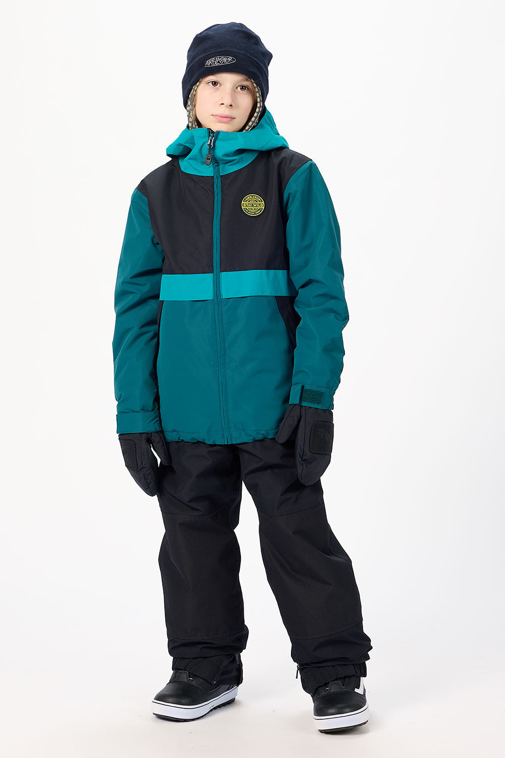 A child wearing a teal and black ski jacket, black snow pants, black gloves, a black beanie, and black sneakers, standing against a plain white studio background.
