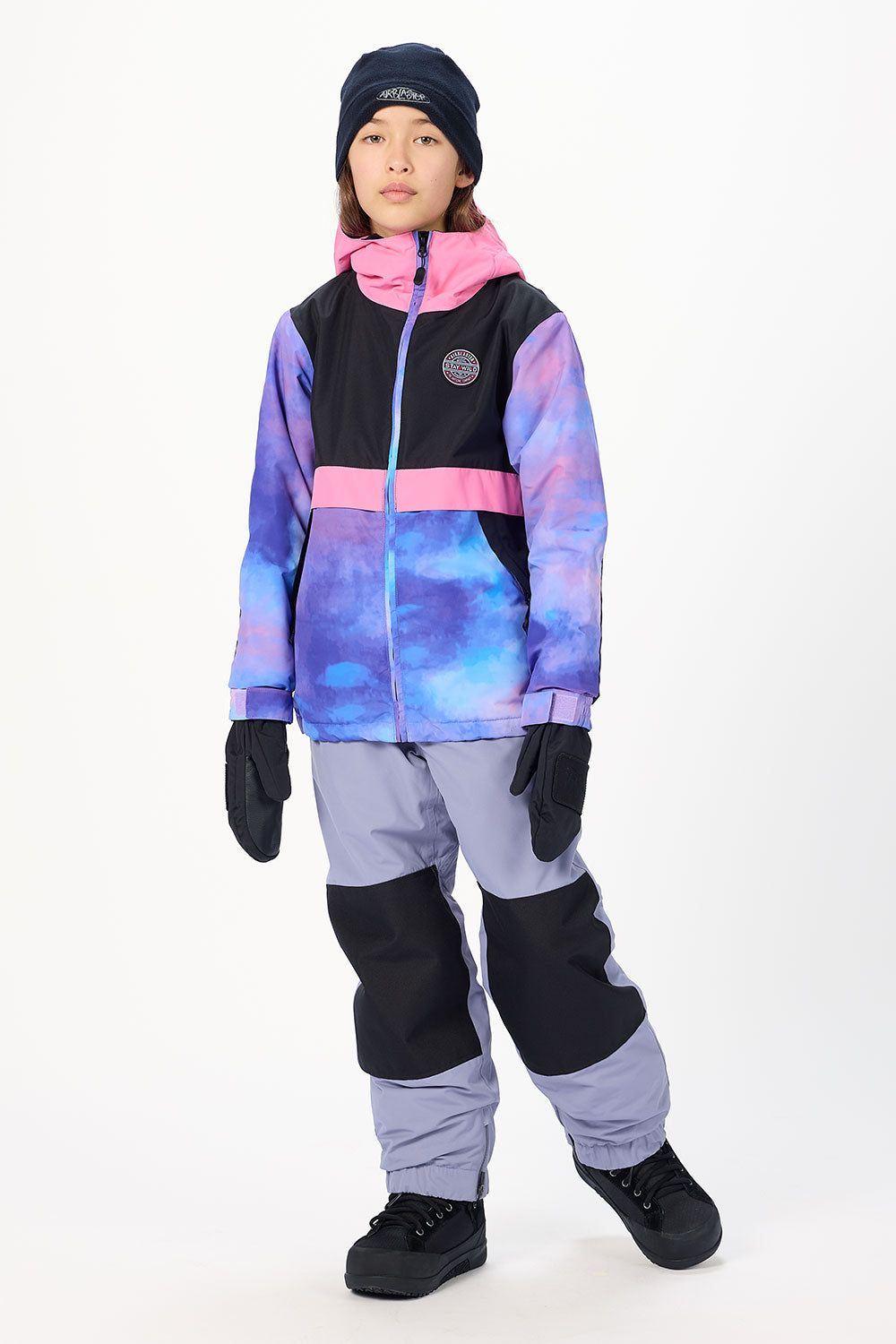 A child model wearing a color-blocked ski jacket with a purple and blue galaxy print, black and pink panels, matching gray ski pants, black gloves, and a black beanie against a white studio background.