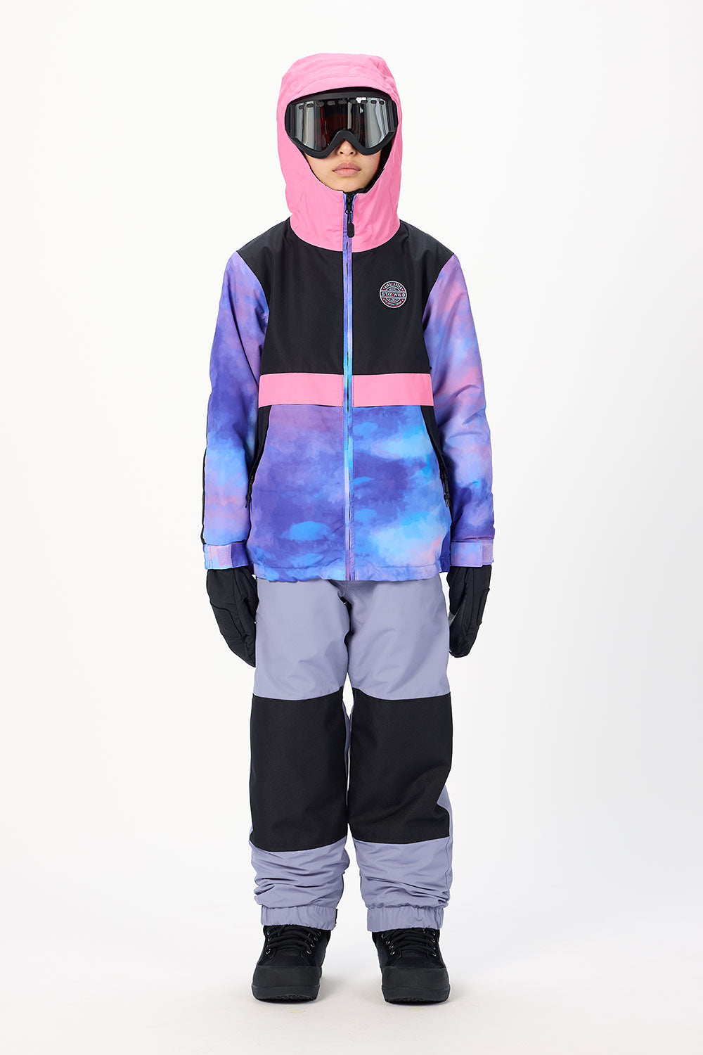 A child modeling a color-blocked winter ski suit with a pink hood, black and purple chest, blue and purple watercolor patterned front, light purple pants, black gloves, and black boots, set against a plain white studio background.