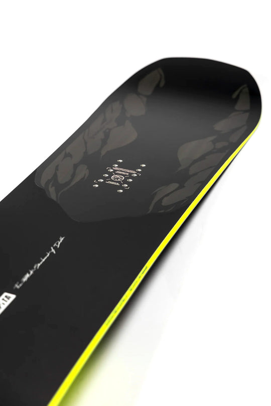Close-up of a snowboard deck showing the base with a black finish, subtle gray graphic pattern, and a bright neon yellow edge stripe on a white background.