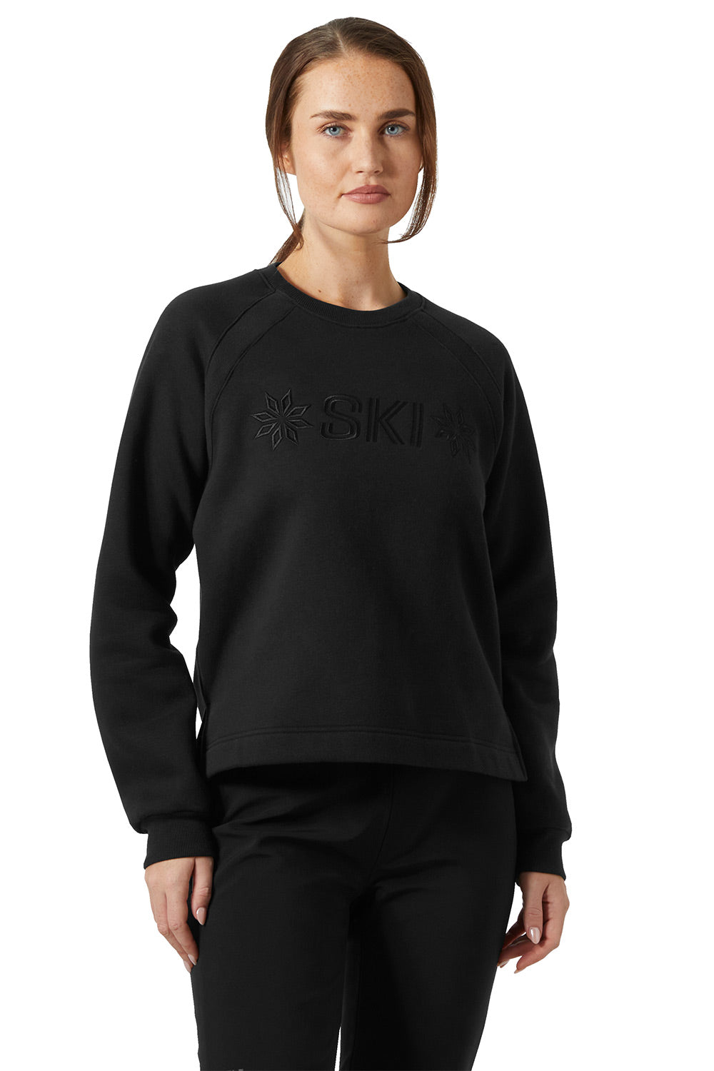 A woman modeling a black crewneck sweatshirt with a tonal black embroidered logo on the chest, paired with black pants, against a plain white background.