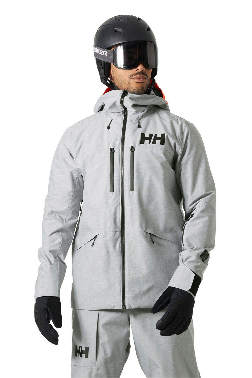 A man wearing a light gray Helly Hansen ski jacket, matching ski pants, black gloves, and a black helmet with goggles, shown against a plain white background.