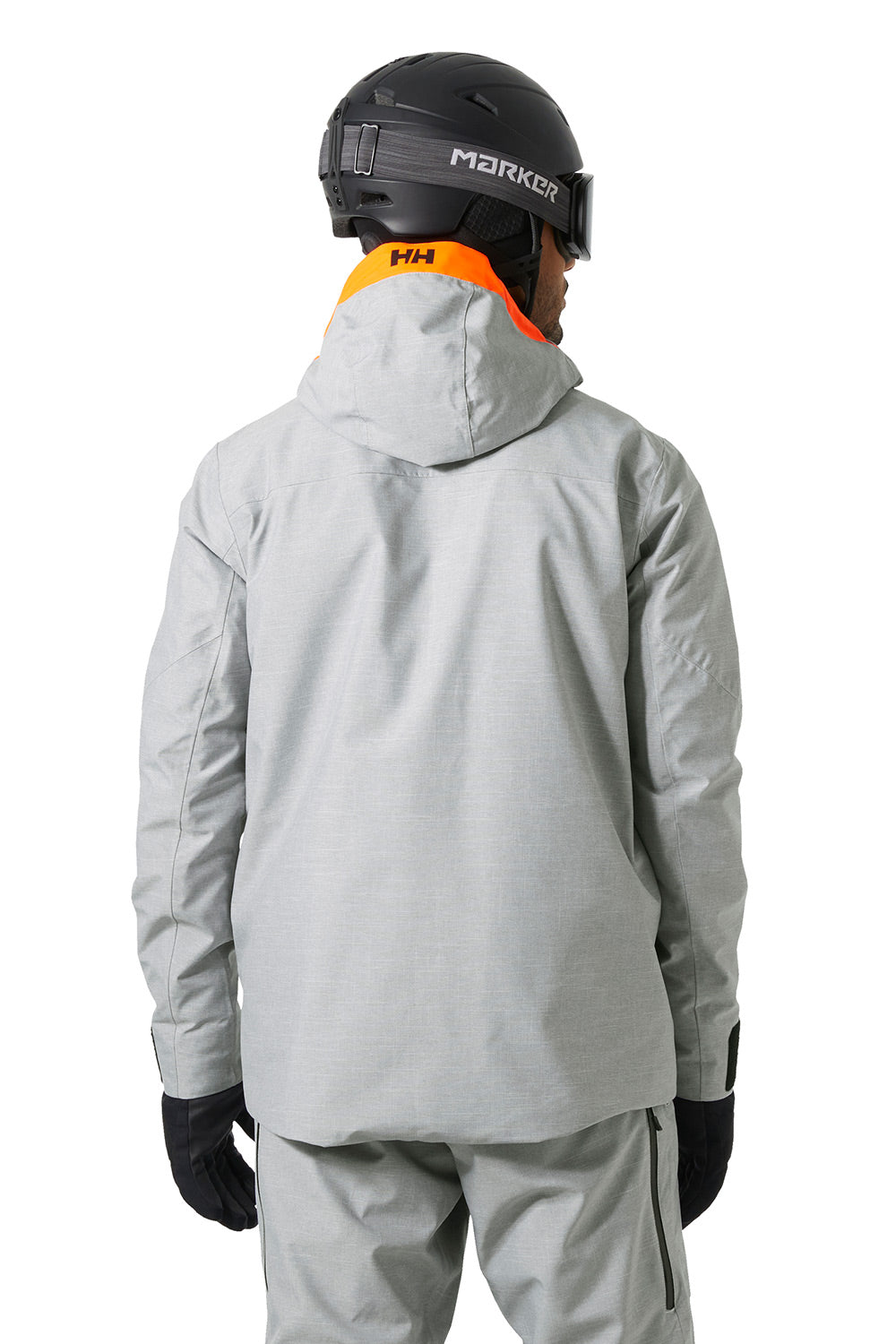 A person shown from the back wearing a light gray hooded ski jacket, matching gray pants, black gloves, a black helmet, and goggles. The jacket has an orange collar lining with an 'Helly Hansen' logo, and the helmet has 'MARKOR' branding.