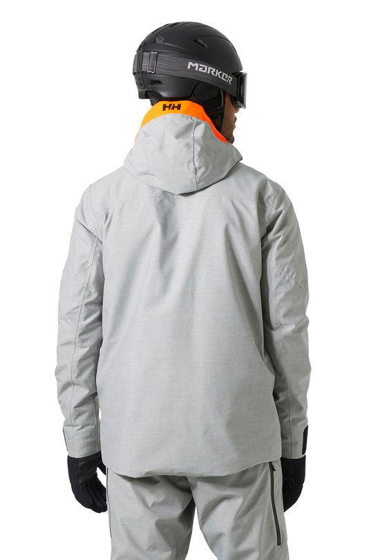 A person shown from the back wearing a light gray hooded ski jacket, matching gray pants, black gloves, a black helmet, and goggles. The jacket has an orange collar lining with an 'Helly Hansen' logo, and the helmet has 'MARKOR' branding.
