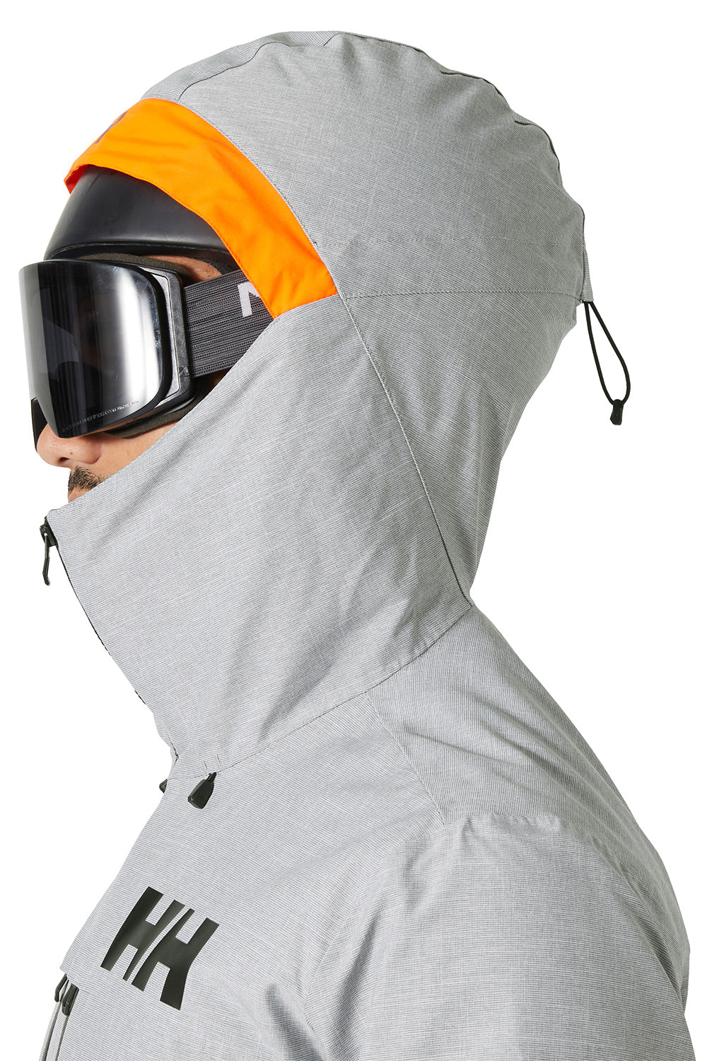 A close-up side profile of a person wearing a light gray Helly Hansen ski jacket with a bright orange hood lining, black goggles, and a black helmet against a white background.