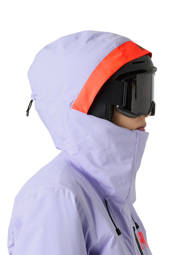 A close-up side profile of a person wearing a light lavender ski jacket with a bright orange accent stripe on the hood, black ski goggles, and a black helmet against a plain white background.