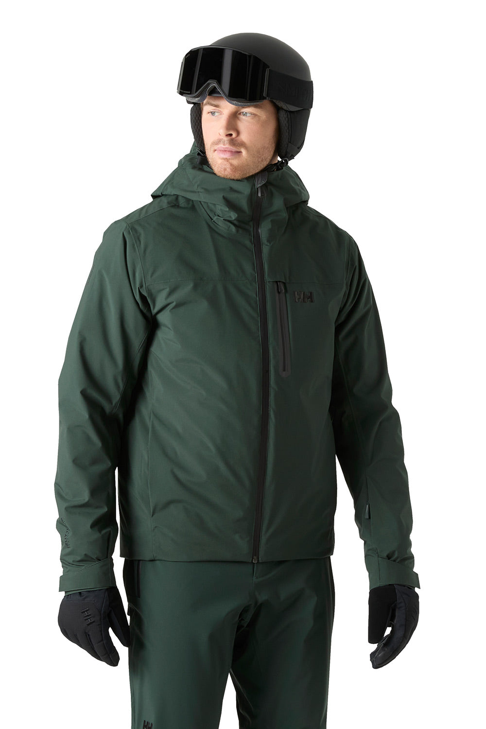A man wearing a dark green ski jacket, matching ski pants, black gloves, a black helmet, and goggles, photographed against a plain white background.