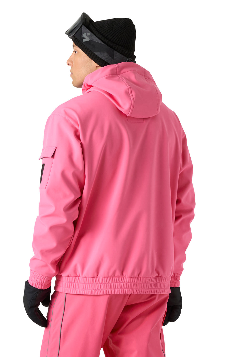 A person shown from the back wearing a bright pink hooded jacket and matching pink pants, with black gloves and a black beanie, against a plain white studio background.
