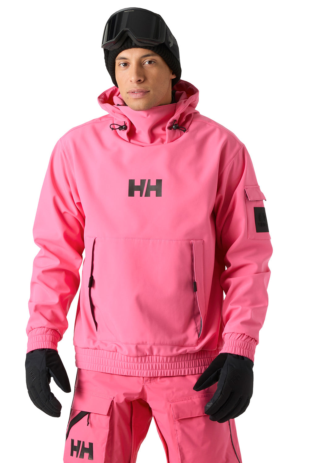 A man wearing a bright pink Helly Hansen snow jacket and matching snow pants, with black gloves, a black beanie, and black ski goggles, photographed against a plain white background.