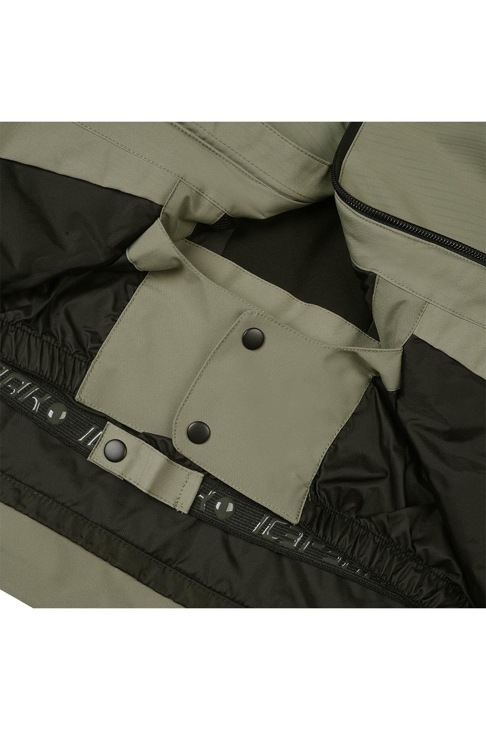 Close-up of a jacket's interior pocket area showing a color-blocked design with olive green fabric and black lining, featuring snap buttons and a black elastic strap with white lettering.