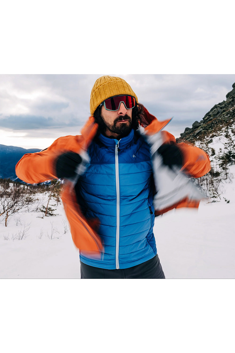 A man in a snowy outdoor setting wearing a bright blue puffer jacket, an orange windbreaker, a yellow knit beanie, and ski goggles.