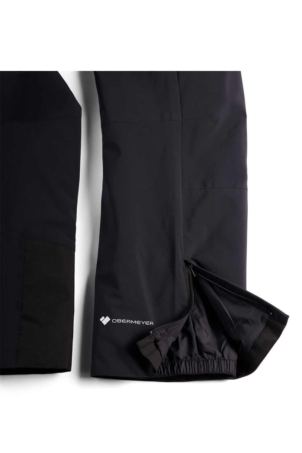 Black technical outdoor pants shown in a close-up studio shot, with one leg folded to reveal an elasticized hem and a small white Obermeyer logo near the lower side.