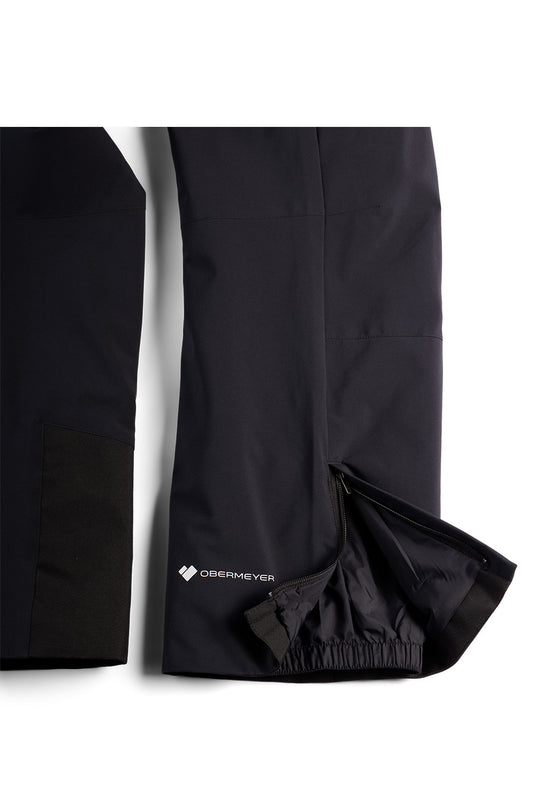Black technical outdoor pants shown in a close-up studio shot, with one leg folded to reveal an elasticized hem and a small white Obermeyer logo near the lower side.