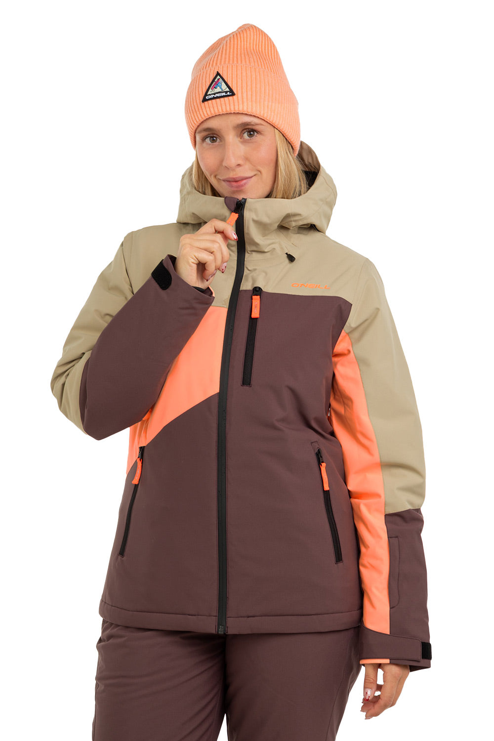 A woman wearing a color-blocked ski jacket in beige, dark brown, and bright orange, paired with a matching orange knit beanie and dark brown ski pants, against a plain white background.