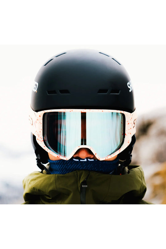 A close-up of a person wearing a matte black ski helmet, light pink patterned ski goggles with mirrored blue lenses, a blue neck gaiter, and an olive green jacket.