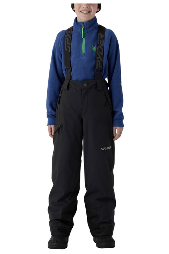 A child model wearing Spyder snow bib overalls in black with a blue quarter-zip fleece underneath, photographed against a plain white studio background.
