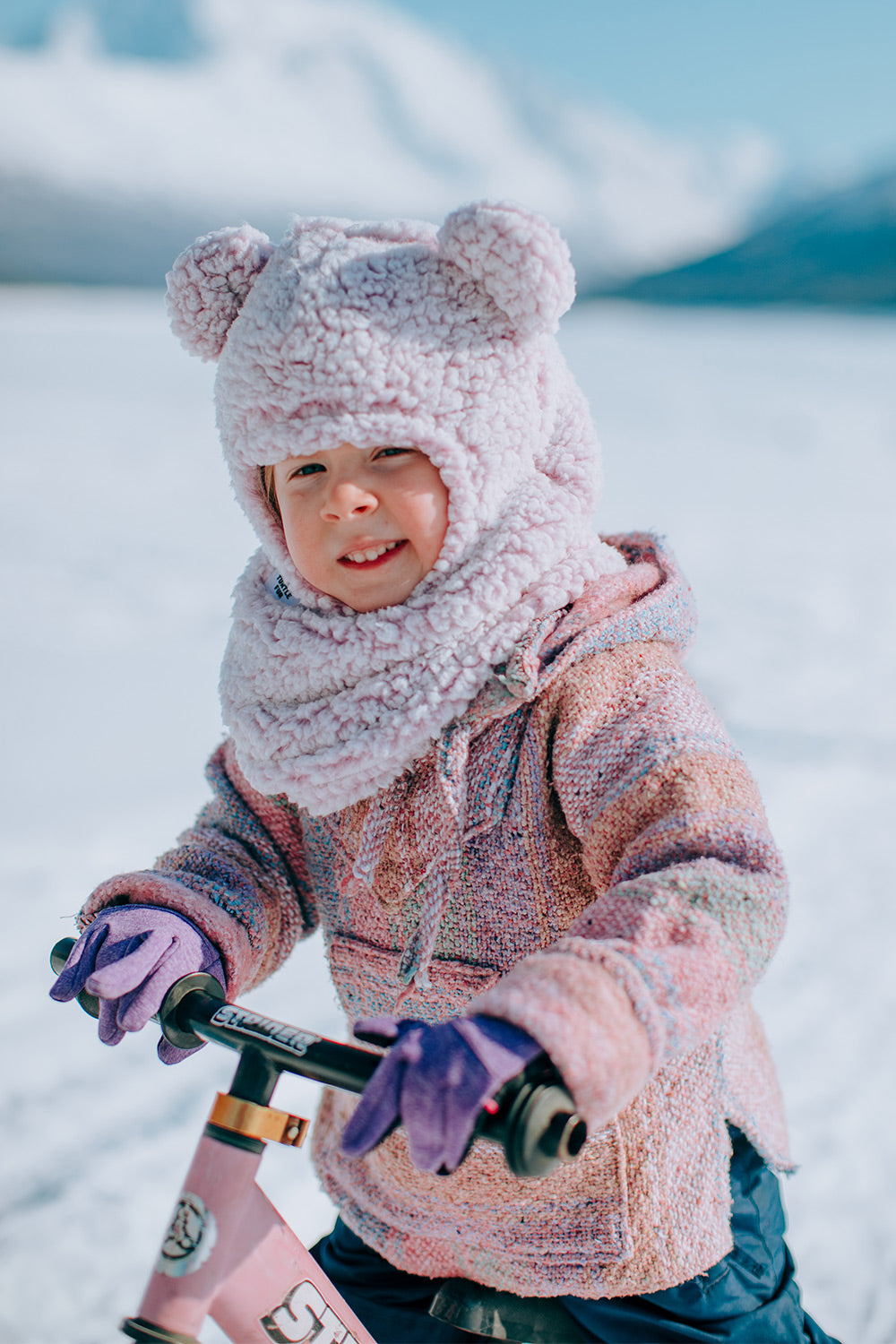 A young child wearing a fuzzy pink and lavender hooded winter jacket, matching scarf, purple gloves, and a bear-ear hat, riding a pink bicycle in a snowy outdoor setting.