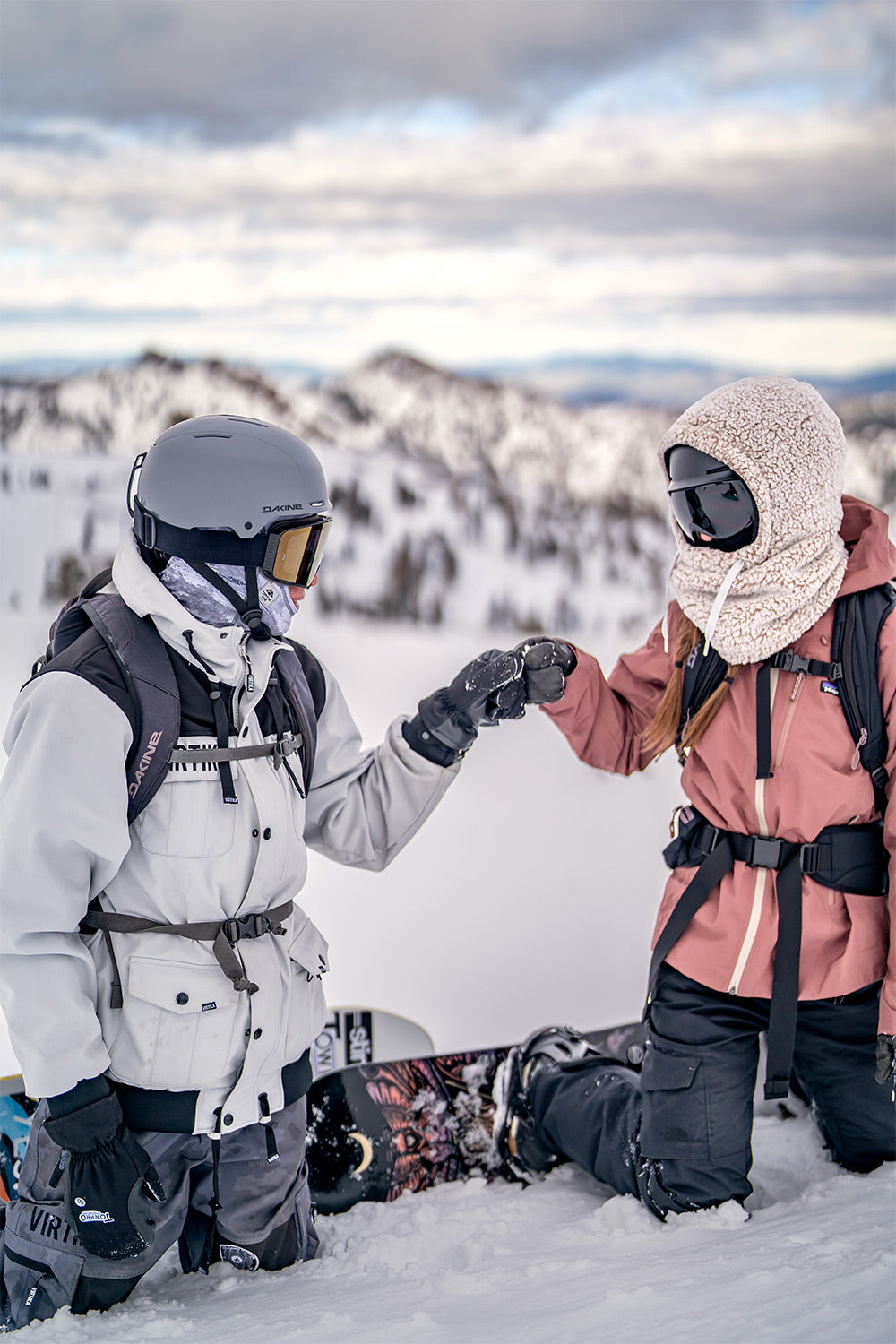 Two snowboarders kneeling in the snow on a mountain, touching gloves in a friendly gesture. One wears a white ski jacket and black helmet, and the other wears a dusty pink hooded ski jacket and black pants.