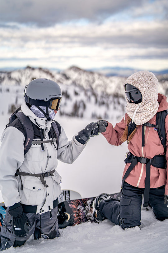 Two snowboarders kneeling in the snow on a mountain, touching gloves in a friendly gesture. One wears a white ski jacket and black helmet, and the other wears a dusty pink hooded ski jacket and black pants.