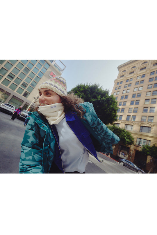 A woman wearing a teal and blue patterned puffer jacket, a white knit beanie, and a white neck warmer, captured in a wide-angle street photograph with city buildings in the background.