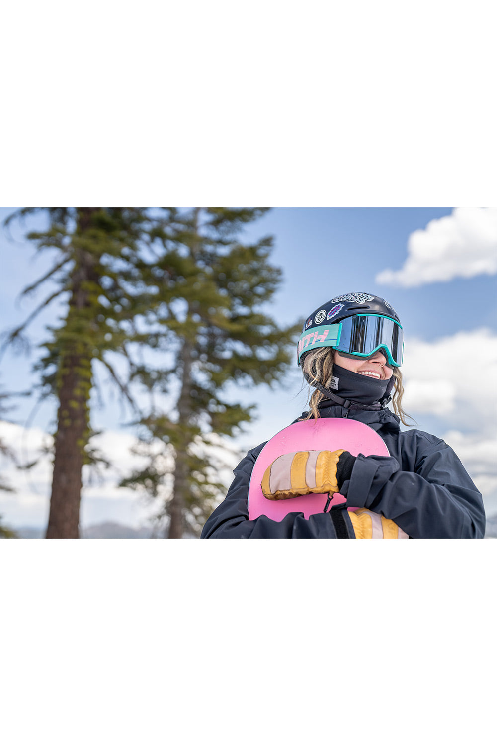 A person outdoors wearing ski gear, including a helmet with mirrored goggles, a black face mask, and gloves, while holding a bright pink snowboard against a background of trees and blue sky.