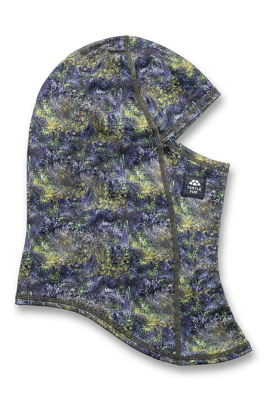 A camouflage balaclava face mask with a hood, featuring a pattern of green, yellow, and dark blue-gray. It has a small black brand patch on the side with the text 'Klimt Peak'.