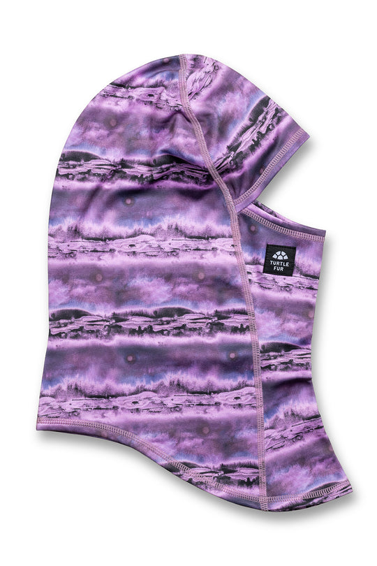A purple and lavender hooded neck warmer or face mask with a scenic mountain landscape print, featuring a small black brand label on the side.