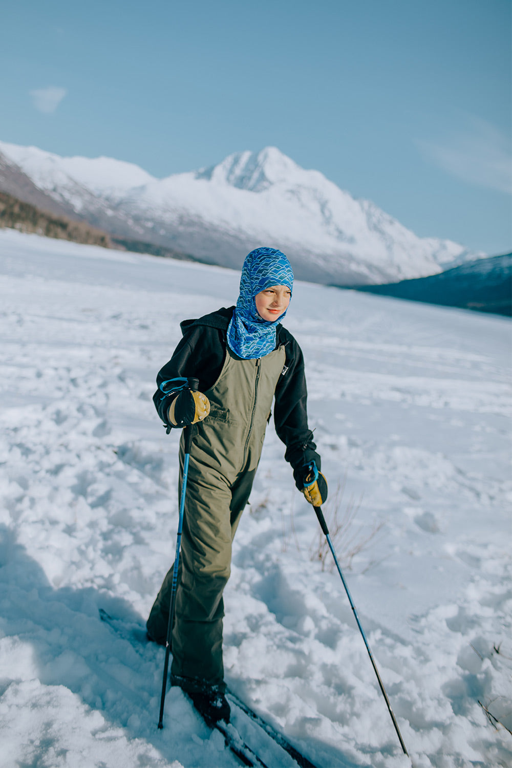A person standing in deep snow on skis, wearing a black and olive green winter ski suit with a blue patterned head covering and holding ski poles, with snow-covered mountains under a blue sky in the background.