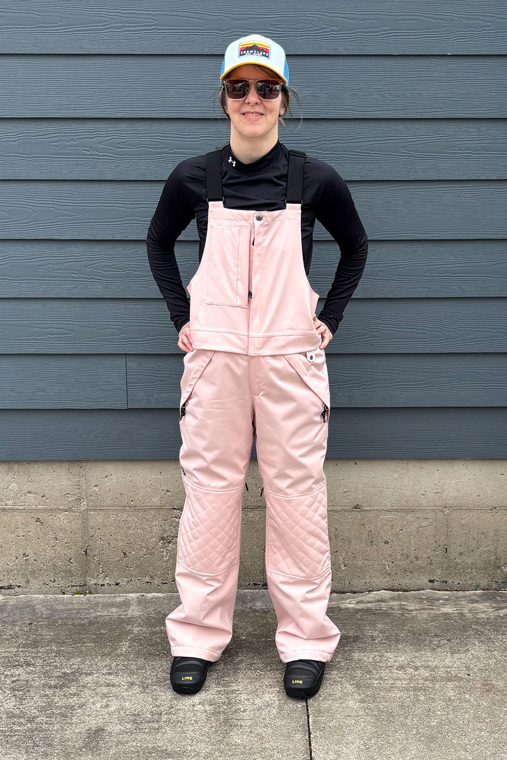 Person wearing pink overalls and a black jacket standing against a blue wall.
