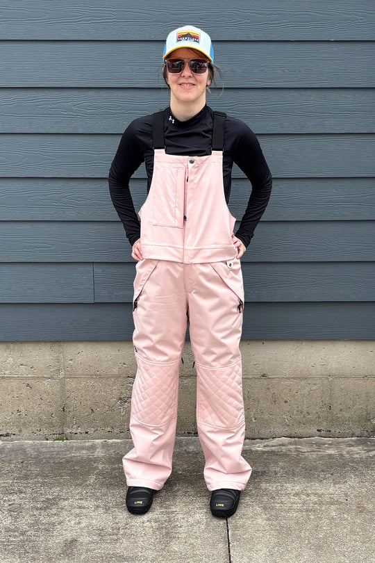 Person wearing pink overalls and a black jacket standing against a blue wall.