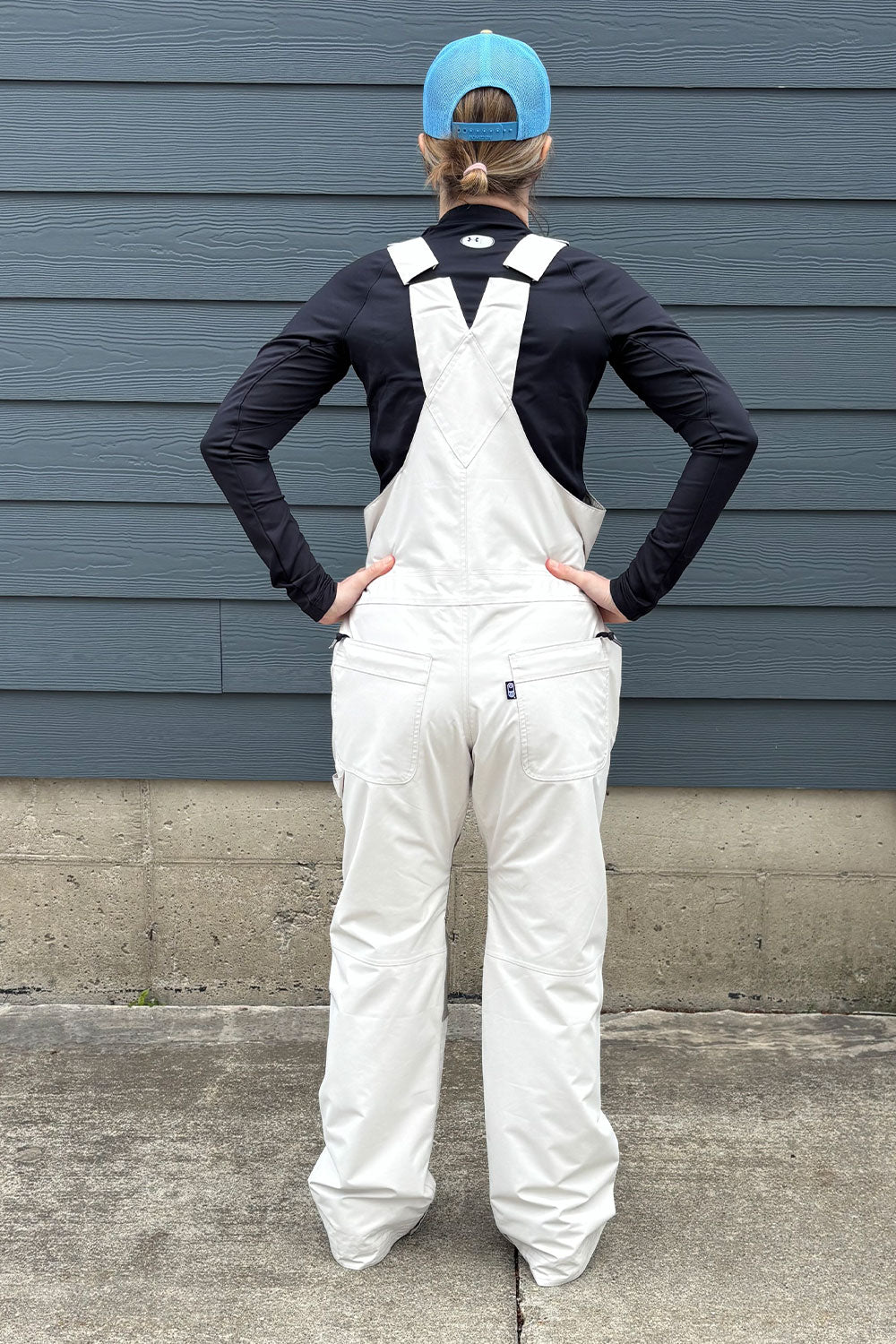 Person wearing white overalls and a blue cap standing against a gray wall.