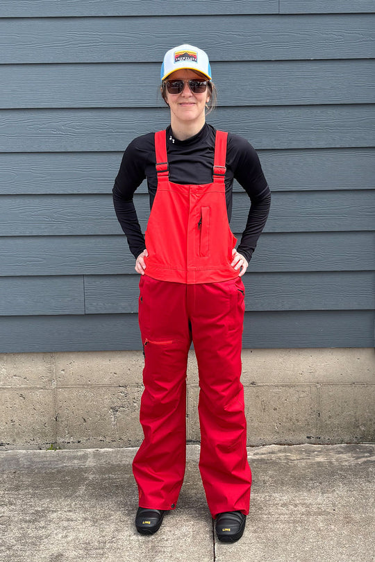 Person wearing red overalls and a black shirt standing against a gray wall.