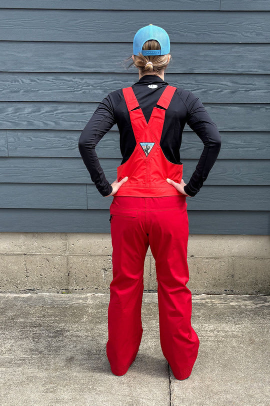 Person wearing a black long-sleeve shirt, red overalls, and a blue cap standing against a gray wall.