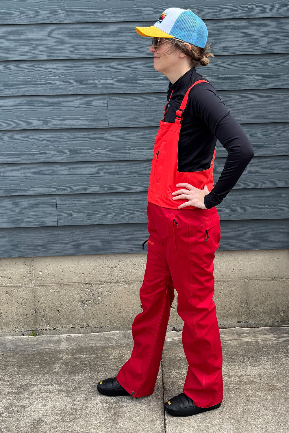 Person wearing red overalls and a colorful cap standing against a gray wall.