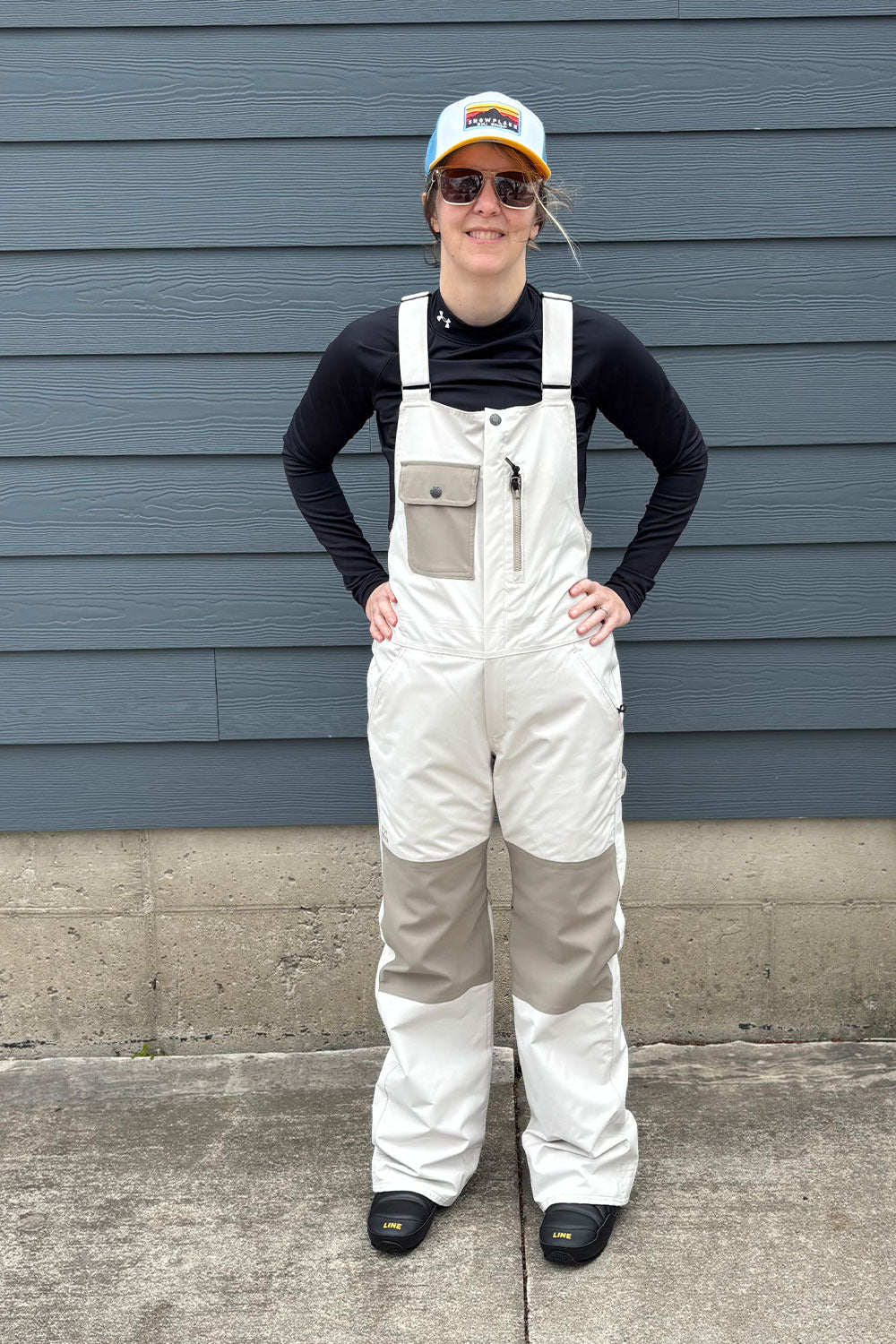 Person wearing white overalls and a black long-sleeve shirt standing against a blue wall.