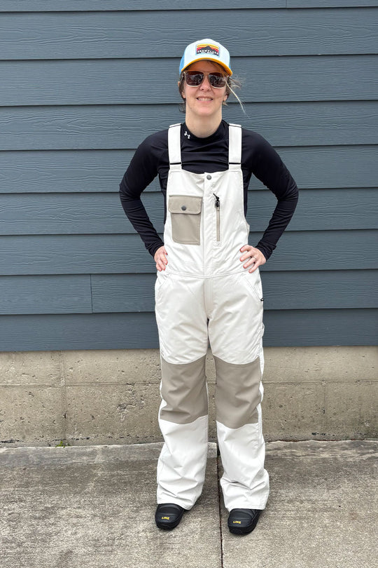 Person wearing white overalls and a black long-sleeve shirt standing against a blue wall.