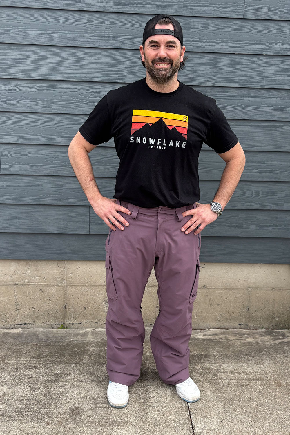 Man wearing a black t-shirt with a graphic design and purple pants standing against a gray wall.