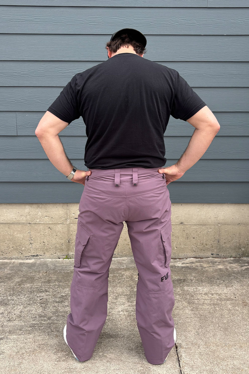 Person wearing a black t-shirt and purple cargo pants standing against a gray wall.